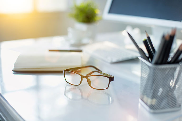 Moderner Schreibtisch mit Brille, Notizbuch, Stiften und Computer bei stimmungsvollem Licht